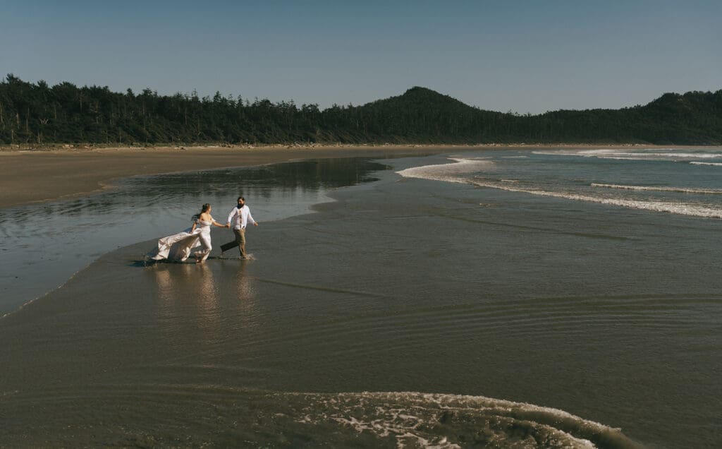 A couple in their wedding attire runs barefoot along the shore at their Tofino Elopement 