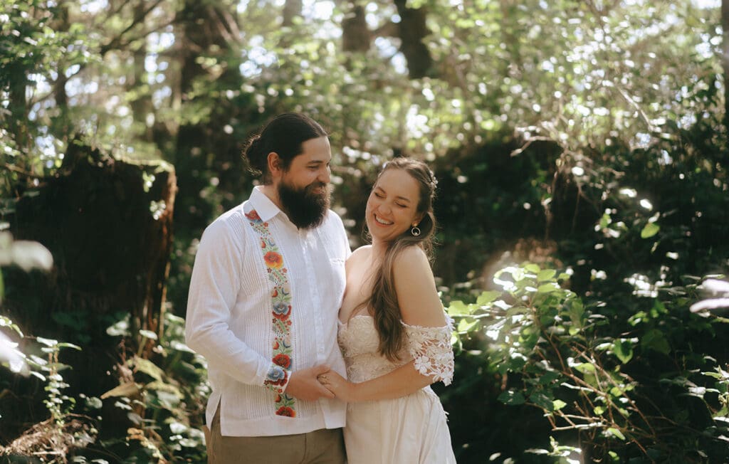 A bride wearing an off the shoulder wedding gown laughs with her groom in the forest at their Tofino elopement 