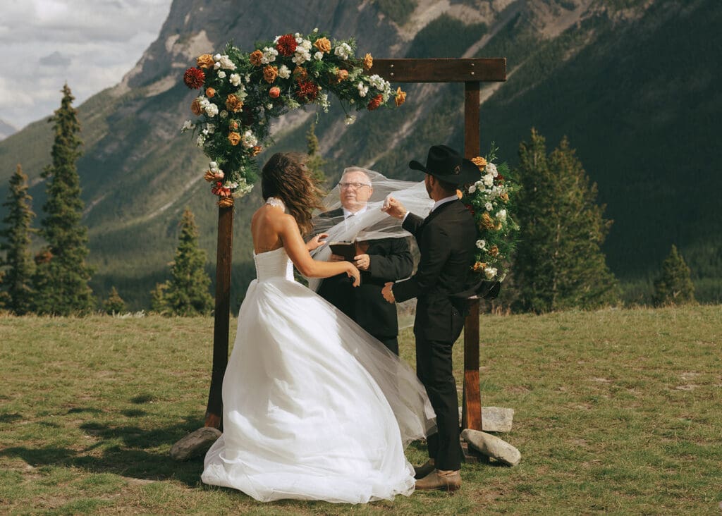 A bride & groom attempt to remove the runaway veil from the officiant during a windy Banff wedding ceremony at Tunnel Mountain Reservoir 