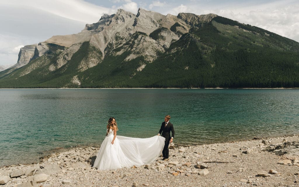 A groom assists the bride with the train of her dress next to the shoreline at Lake Moraine before their Tunnel Mountain Reservoir wedding ceremony in Banff