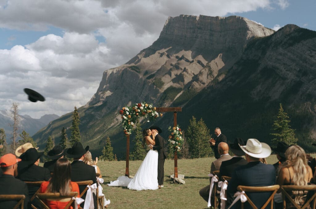 Newlyweds share a kiss beneath a wooden arch at their Banff wedding ceremony at Tunnel Mountain Reservoir