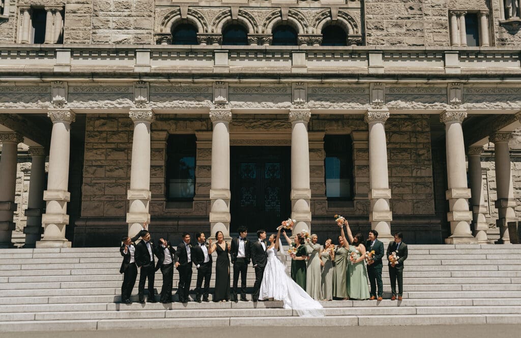 A bride and groom along with their large wedding party pose for a bridal portrait on the steps of parliament house victoria bc