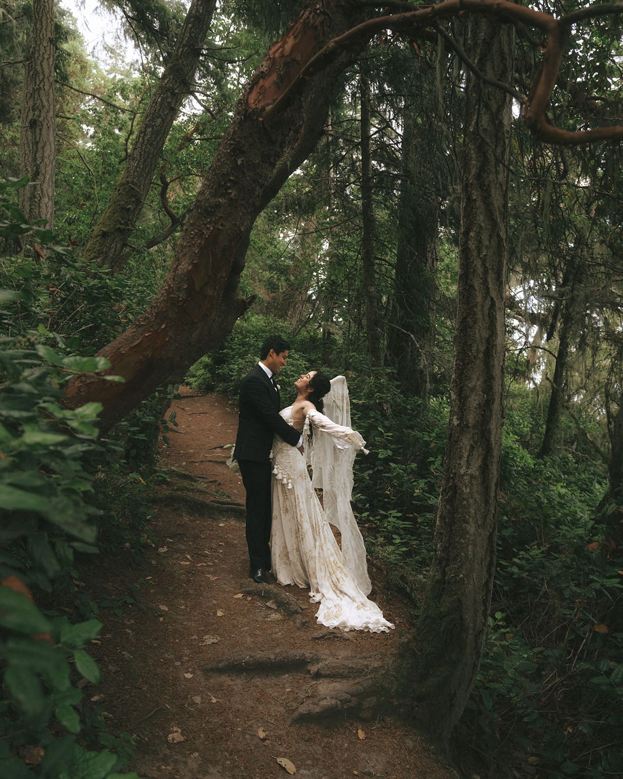 groom hugging bride who has her arms up showing off the long sleeves on her wedding dress while standing in forest