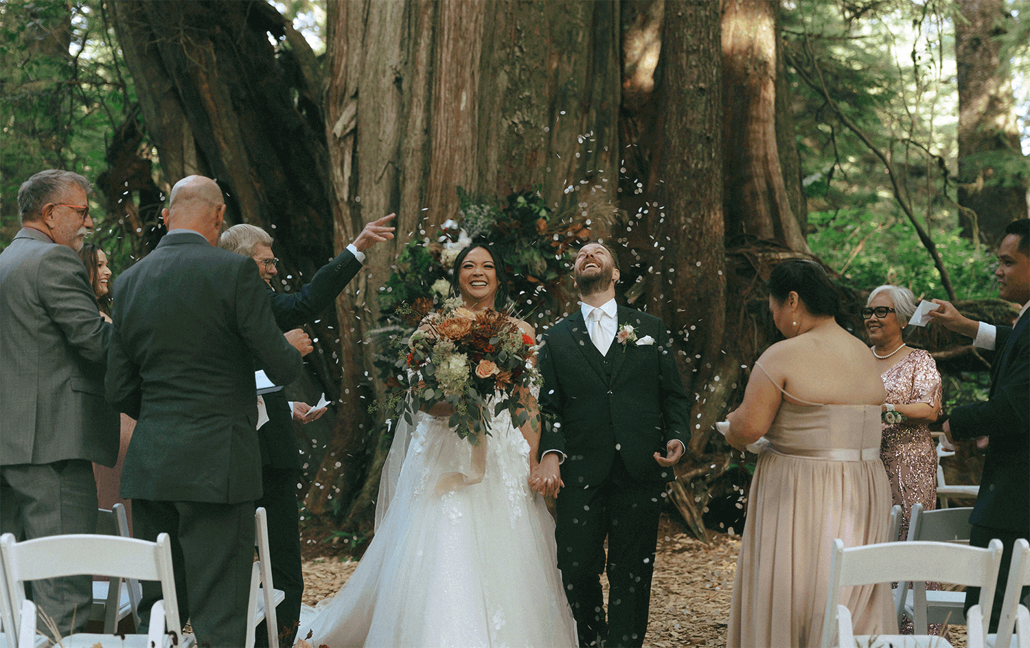 bride and groom holding hands and smiling as they exit wedding ceremony in the forest while guests throw flower petals