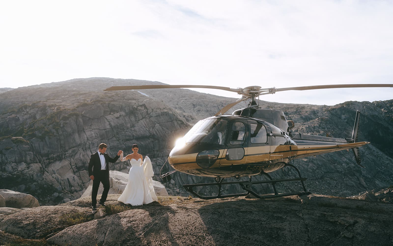 groom holding bride's hand and helping her walk on mountain with a helicopter next to them